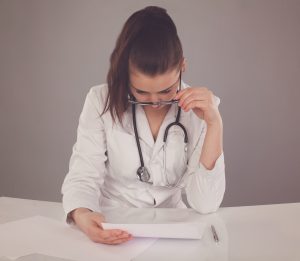 Nurse in white robe and glasses and stethoscope is checking her report and thinking about it at the white table against of grey background